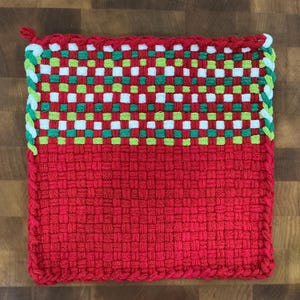 May include: A square, woven potholder with a red base and a checkered pattern of white, green, and red in the upper section. The edges are trimmed with white and green yarn. A small loop is attached at the top corner.