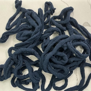 May include: A tangled pile of navy blue knitted rope. The rope is thick and appears to be made of a soft, textured material. The image is taken from a top-down perspective, showing the rope against a white surface.