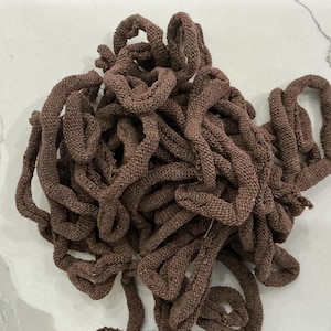 May include: A pile of brown knitted fabric, possibly yarn or cord, with a textured appearance. The material is a warm, earthy tone, and the strands are looped and intertwined, creating a dense, tactile display. The fabric appears to be handmade.