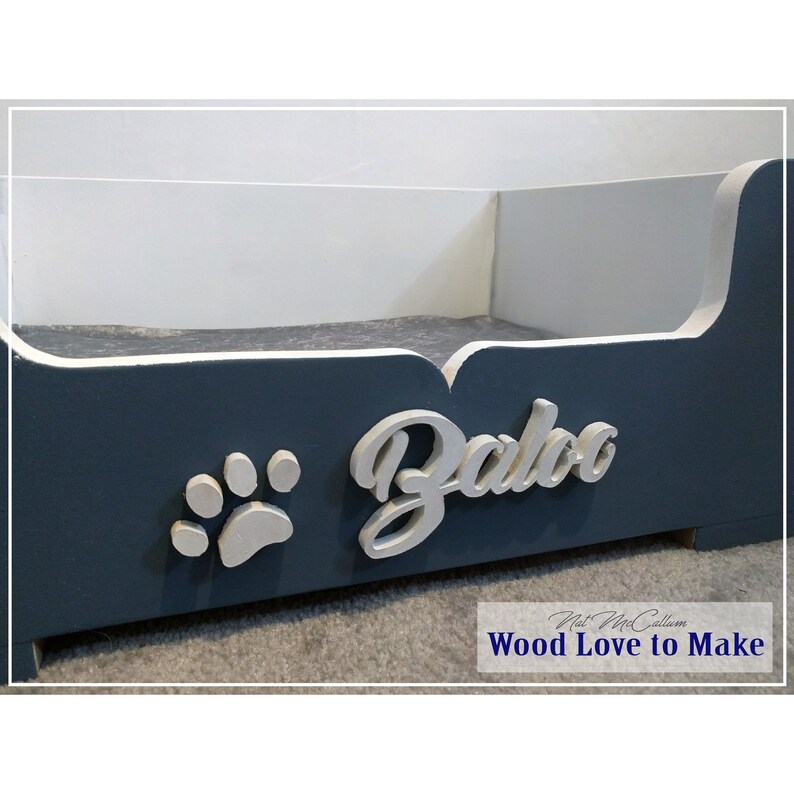 Puede incluir: Una cama de perro de madera azul con una huella de pata blanca y el nombre "Baloo" escrito en blanco en la parte delantera. La cama est&aacute; hecha por "Wood Love to Make".