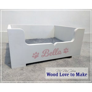 Puede incluir: Cama de perro de madera blanca con un coj&iacute;n gris y el nombre "Bella" escrito en rosa con huellas de patas a cada lado.