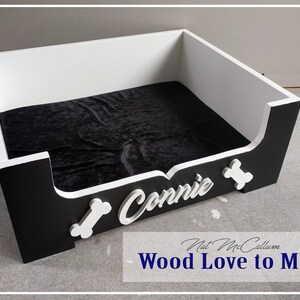 Puede incluir: Una cama para perros negra y blanca con una placa de nombre blanca que dice "Connie" y dos formas de hueso blancas. La cama tiene un forro de terciopelo negro.