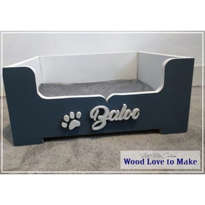 Puede incluir: Una cama para perro de madera azul y blanca con un coj&iacute;n gris. La cama tiene una huella de pata y el nombre "Baloo" escrito en la parte delantera.
