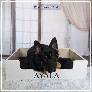 Puede incluir: Una cama de perro de madera blanca con un coj&iacute;n negro y el nombre "AYALA" grabado en la parte delantera. Un perro pastor alem&aacute;n negro y marr&oacute;n est&aacute; acostado en la cama.