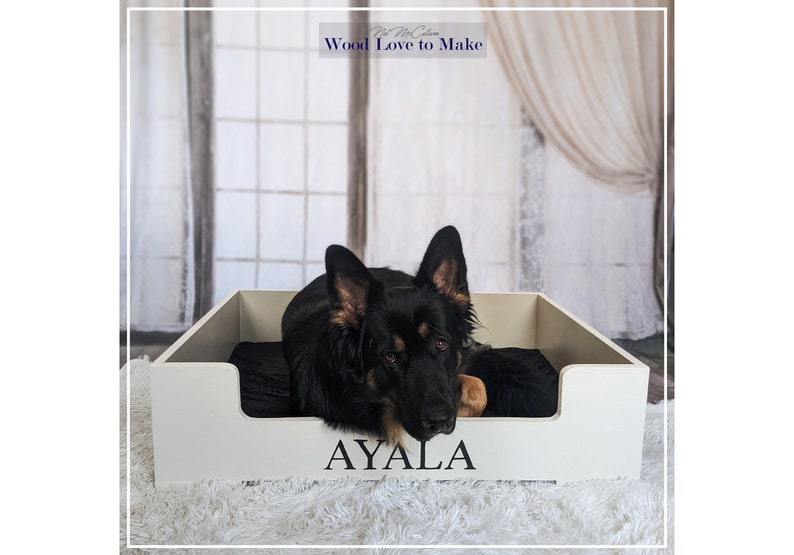 Puede incluir: Una cama de perro de madera blanca con un coj&iacute;n negro y el nombre "AYALA" grabado en la parte delantera. Un perro pastor alem&aacute;n negro y marr&oacute;n est&aacute; acostado en la cama.