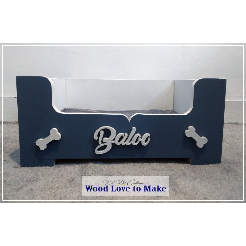 Puede incluir: Una cama para perro de madera azul y blanca con el nombre "Baloo" en la parte delantera. La cama tiene dos recortes en forma de hueso en los lados.