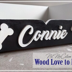 Puede incluir: Cama de perro de madera negra con detalles de huesos blancos y el nombre "Connie" escrito en blanco en la parte delantera.