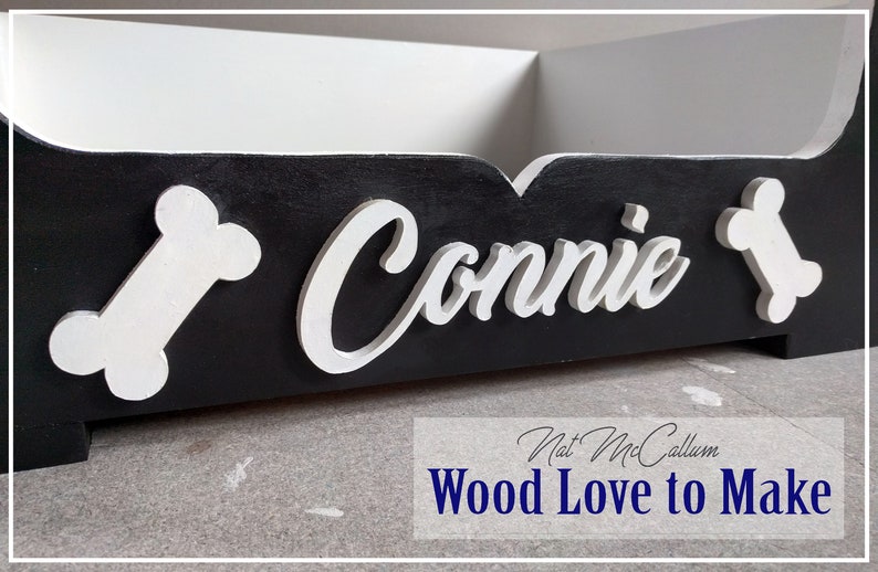 Puede incluir: Cama de perro de madera negra con detalles de huesos blancos y el nombre "Connie" escrito en blanco en la parte delantera.