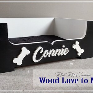 Puede incluir: Cama de perro de madera negra y blanca con el nombre "Connie" y formas de hueso en la parte delantera.