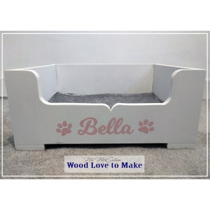 Puede incluir: Cama de perro de madera blanca con un coj&iacute;n gris y el nombre "Bella" en letras rosas con huellas de patas a cada lado. La cama est&aacute; sobre una alfombra gris.