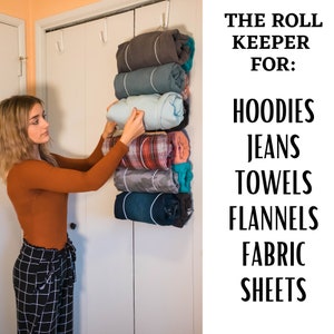 May include: A woman is organizing rolled up clothing using a hanging rack. The rack is labeled "The Roll Keeper for: Hoodies, Jeans, Towels, Flannels, Fabric Sheets".