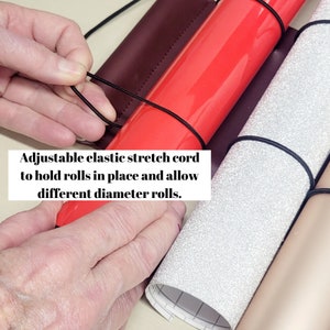 May include: A close-up of a craft storage organizer with adjustable elastic cords to hold rolls of different sizes. The organizer has a burgundy, red, silver, and gold fabric.