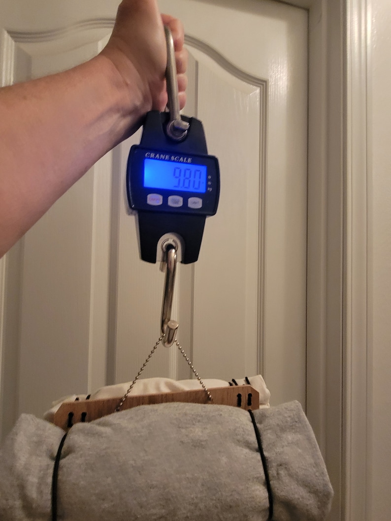 May include: A black digital crane scale with a hook hanging a gray and white blanket. The scale reads 9.80.