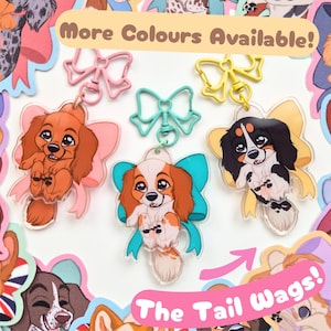 May include: Three acrylic keychains featuring cartoon spaniel puppies with large eyes, each attached to a matching colored bow-shaped clasp. The puppies are brown, white and brown, and black and white. Text reads More Colours Available! and The Tail Wags!