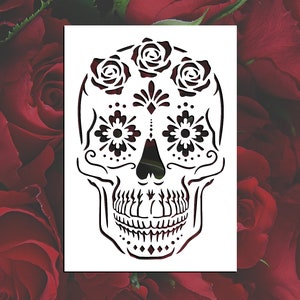 May include: A white stencil of a sugar skull with floral designs. The skull has three roses on top, and intricate floral patterns around the eyes and mouth.