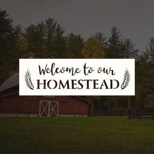 May include: A white sign with black text and a wheat stalk design on each side reads "Welcome to our HOMESTEAD". A red barn with a white door and two windows is in the background.