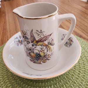 May include: White ceramic pitcher and saucer set with gold trim. The pitcher features a colorful bird and fruit basket design. The saucer and pitcher are on a green placemat.