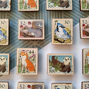 May include: Wooden postage stamp magnets featuring various animals, including a squirrel, blue jay, fox, otter, and Shiba Inu. Each magnet has a unique design with a vintage aesthetic, including the text "AMSTERDAM" and a price.