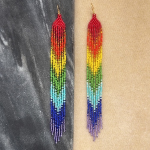 May include: A pair of long, rainbow-colored beaded earrings. The earrings are made with small, colorful beads that are strung together in a chevron pattern. The earrings are about 3 inches long and have a gold hook closure.