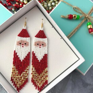 May include: A pair of red, white, and gold beaded earrings featuring a Santa Claus face. The earrings are displayed in a white gift box with a turquoise lid.