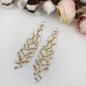 Puede incluir: Un par de aretes largos de cuentas blancas con un patr&oacute;n geom&eacute;trico dorado. Los aretes cuelgan y tienen un dise&ntilde;o de flecos.
