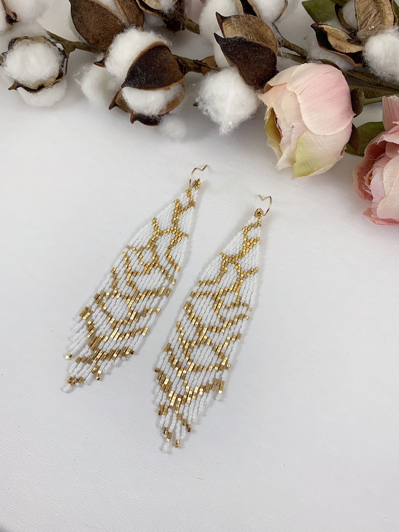 Puede incluir: Un par de aretes largos de cuentas blancas con un patr&oacute;n geom&eacute;trico dorado. Los aretes cuelgan y tienen un dise&ntilde;o de flecos.