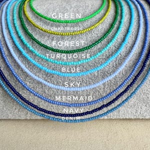 May include: A collection of beaded necklaces in various shades of blue and green. The necklaces are arranged in a layered display, with each necklace showcasing a different color, including green, chartreuse, forest, turquoise, blue, sky, mermaid, navy, and denim. Text labels identify each color.