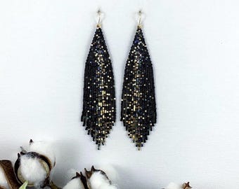 May include: A pair of long, beaded earrings. The earrings are black with gold and silver accents, and they dangle from gold-colored hooks. The earrings are teardrop-shaped and have a fringe at the bottom. The background is white.