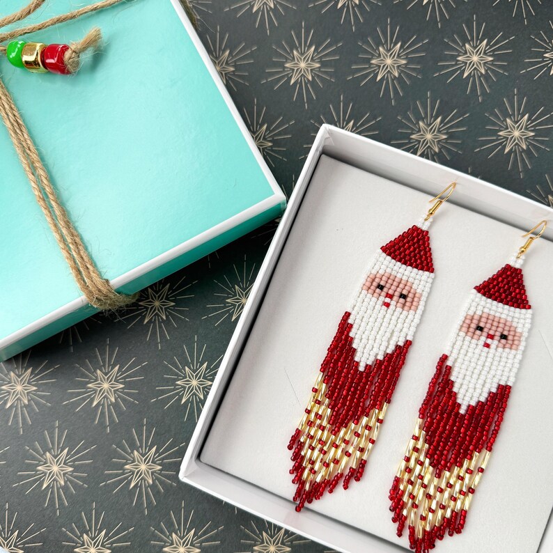 May include: A pair of red and white beaded Santa Claus earrings with gold accents. The earrings are in a white gift box.