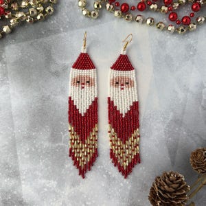 May include: Red and white beaded earrings with a Santa Claus design. The earrings are long and dangle, with gold beads at the bottom.