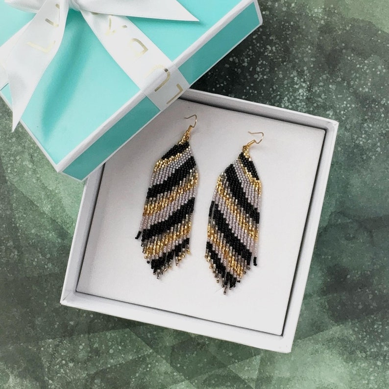 May include: A pair of black, gold, and silver beaded earrings with a fringe design. The earrings are displayed in a white gift box.