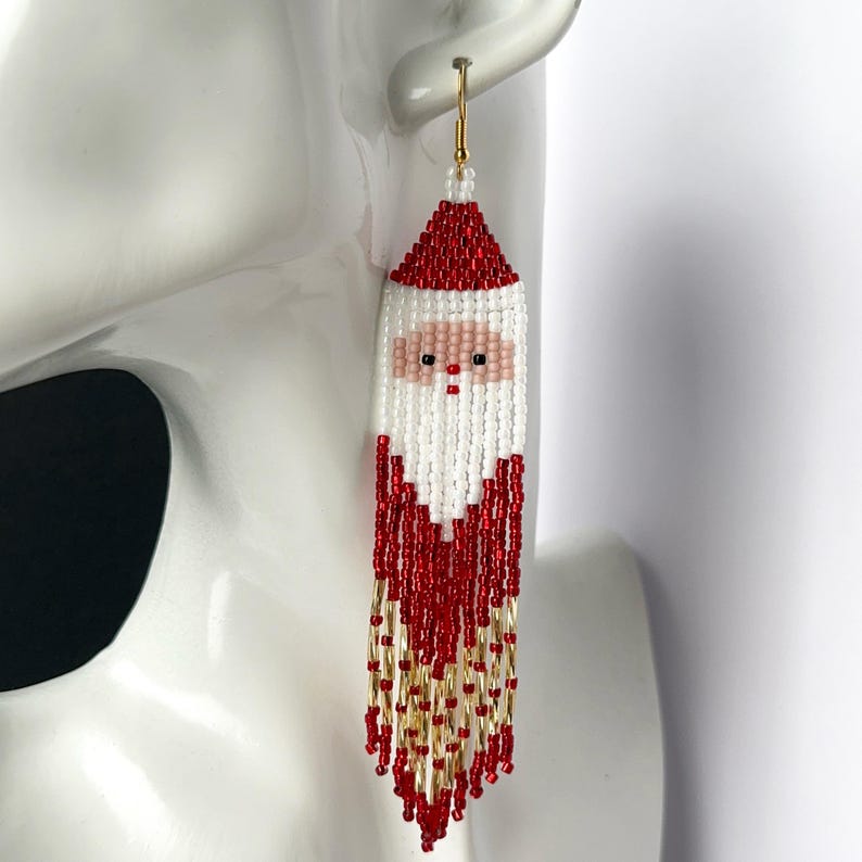 May include: Red and white beaded earrings with a Santa Claus face design. The earrings have a gold hook and a long fringe of red and gold beads.