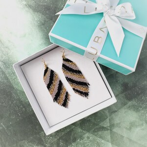 May include: A pair of long, beaded earrings in a black, gold, and white geometric pattern. The earrings are displayed in a white gift box with a turquoise lid.
