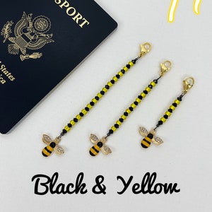 May include: Three black and yellow beaded chains with a gold bee charm at the end. The chains are hanging from a white surface. The text "Black & Yellow" is written below the chains.