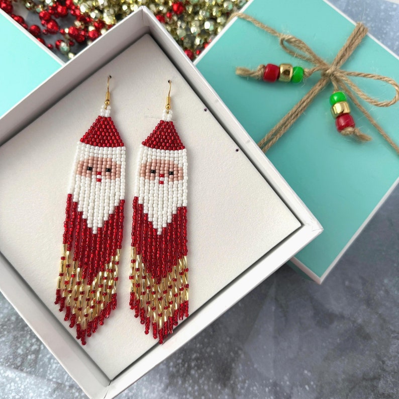 May include: A pair of red, white, and gold beaded earrings featuring a Santa Claus face. The earrings are displayed in a white gift box with a turquoise lid.