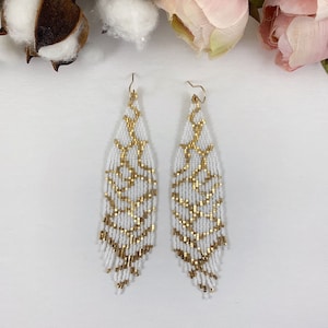 Puede incluir: Un par de aretes largos de cuentas blancas con detalles dorados. Los aretes tienen forma de l&aacute;grima y un dise&ntilde;o de flecos.
