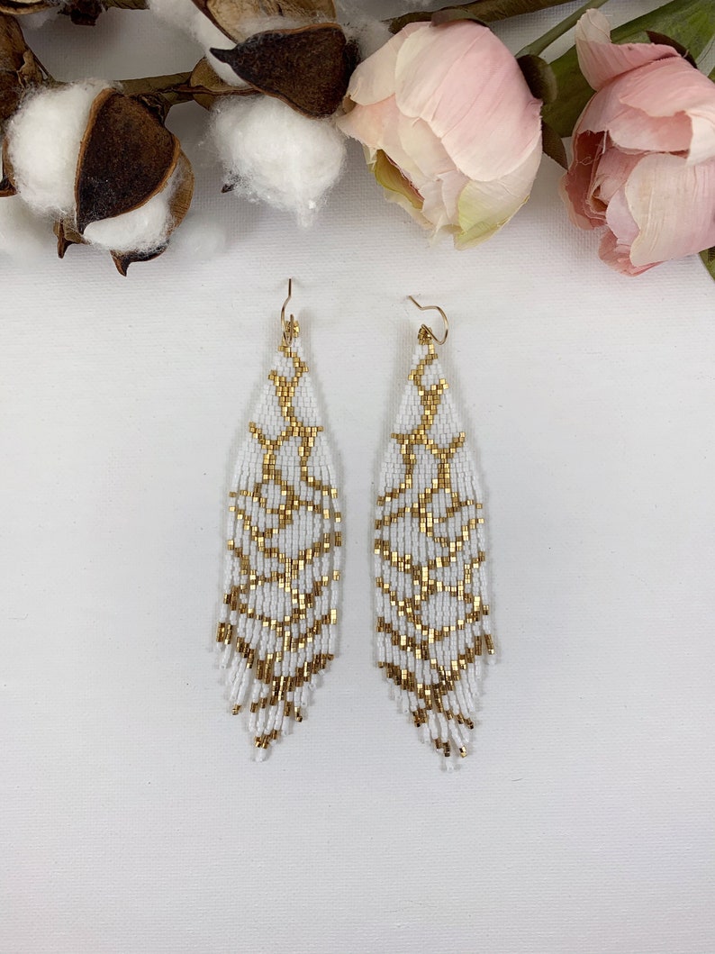 Puede incluir: Un par de aretes largos de cuentas blancas con detalles dorados. Los aretes tienen forma de l&aacute;grima y un dise&ntilde;o de flecos.