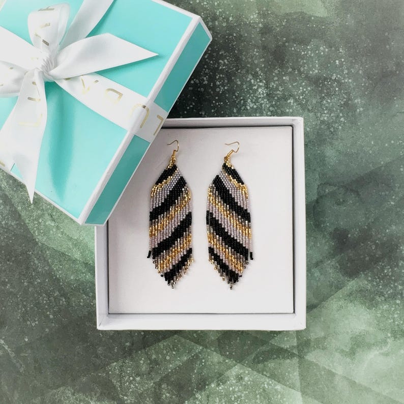 May include: A pair of long, beaded earrings in a black, gold, and silver color scheme. The earrings are in a gift box.