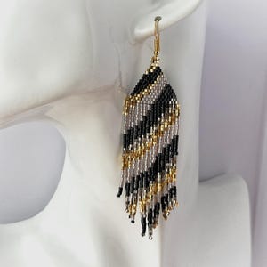 May include: A pair of long, beaded dangle earrings with a black, gold, and silver geometric pattern. The earrings are made with small, seed beads and have a tassel-like fringe.