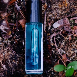 May include: A small blue glass bottle with a black cap and a white label. The bottle is filled with a clear liquid and is sitting on a bed of brown leaves and moss.