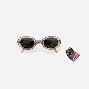 May include: Oval-shaped sunglasses with a textured, light-colored frame and dark lenses. The sunglasses have a silver-colored bridge and temples. A tag is attached to the sunglasses.