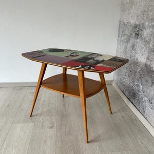 Könnte beinhalten: Ein Beistelltisch im Mid-Century-Stil mit einer rechteckigen Tischplatte mit einer Collage aus roten, grünen und schwarzen Bildern. Der Tisch hat ein unteres Regal und schlanke, sich verjüngende Beine. Der Tisch ist ca. 61 cm lang, 41 cm breit und 51 cm hoch.