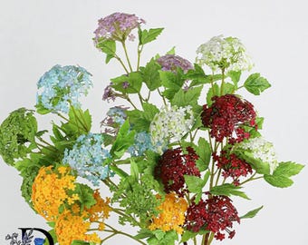 May include: A bouquet of artificial flowers featuring clusters of small blossoms in shades of yellow, light blue, white, purple, and deep red. The arrangement includes green serrated leaves on thin stems. A logo with the letter D and a blue rose is visible.