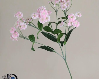 May include: A single stem of artificial flowers featuring multiple light pink blossoms with five petals each. The stem has thin green branches and several pointed, dark green leaves. A logo with the letter D and a blue rose is in the corner.
