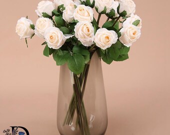 May include: A bouquet of artificial cream-colored roses with green leaves arranged in a tall, translucent, smoke-gray glass vase. The flowers feature layered petals and are supported by thin, dark green stems that extend into the clear glass vessel.