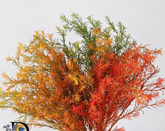 May include: A bundle of artificial foliage featuring thin, needle-like leaves in a gradient of colors ranging from deep forest green at the top to vibrant orange and bright red at the base, with a logo featuring a blue rose and the text DOZEN.