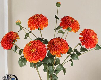 May include: A bouquet of artificial dahlia flowers featuring vibrant orange and yellow petals arranged on long green stems with dark green leaves. The flowers display a dense, layered petal structure with several buds at different stages.
