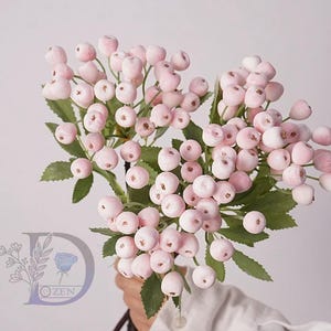 May include: A bunch of artificial pink berries with green leaves. The berries are grouped on stems, giving a full and textured look. The leaves are a deep green, contrasting with the soft pink berries. The bouquet is held in a person's hand.