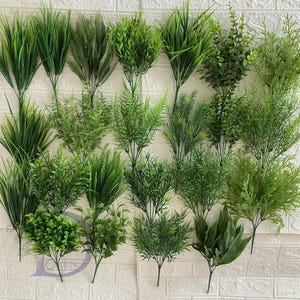 Può includere: Un assortimento di piante verdi artificiali, tra cui vari tipi di erba e steli fogliosi. Le piante sono disposte contro un muro di mattoni bianchi, mostrando una varietà di trame e tonalità di verde, perfette per l'arredamento della casa o progetti di artigianato.