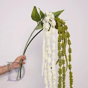 Puede incluir: Tallos florales colgantes artificiales en blanco y verde, sostenidos por una mano. Los tallos presentan flores en cascada y hojas verdes sobre un fondo blanco.